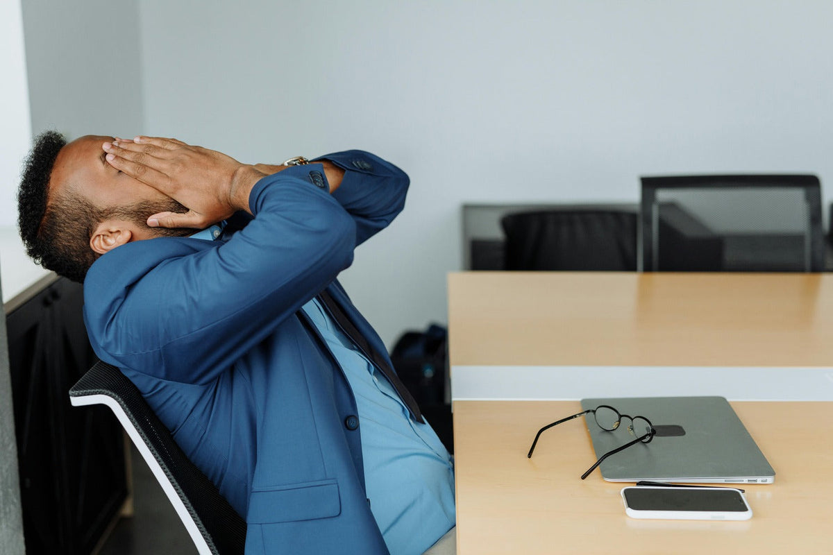 A man holding his head looking stressed with laptop and glasses on desk